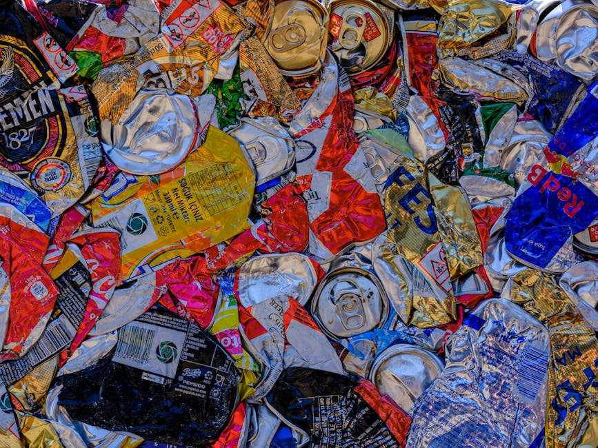 Close-up image of a large pile of crushed and wrinkled aluminum cans and metal beverage containers, predominantly in silver, gold, red, blue, and black colors, with visible branding, barcodes, and labels. The cans are randomly scattered and overlapping, showing various textures from smooth and shiny to slightly dents and creases, indicating they have been flattened for recycling or disposal. The scene appears to be set outdoors or on a flat surface, with no background elements visible. This collection of waste suggests an example of alternative waste handling, which Rubbish Collection Haringey could include in their rubbish removal services, especially for events where numerous cans and metals are disposed of efficiently through on-site clearance or private collection methods.