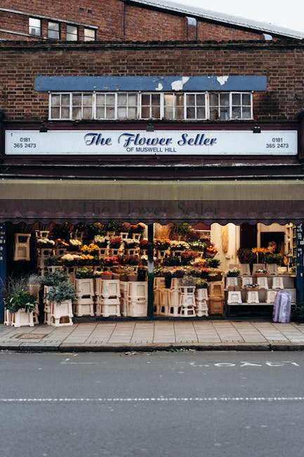 The image shows the storefront of a flower shop called 'The Flower Seller of Muswell Hill,' situated on a pavement beside a road. The shop has a dark pink or maroon awning with the store's name displayed in white cursive lettering, and several flower arrangements and potted plants are visible through the open front window and on outdoor displays. Above the shop, there is a blue sign with white text indicating the business name and contact numbers, mounted on a brick building with a small section of weathered white paint or damage near the roofline. The shop appears busy with various floral displays arranged on tables, shelves, and stands, and some plants are in white or beige containers. The overall scene captures a typical urban commercial frontage with a focus on floral retail, and the setting suggests potential waste or rubbish removal activities related to maintaining the premises, consistent with private waste handling services possibly offered by companies like Rubbish Collection Haringey.