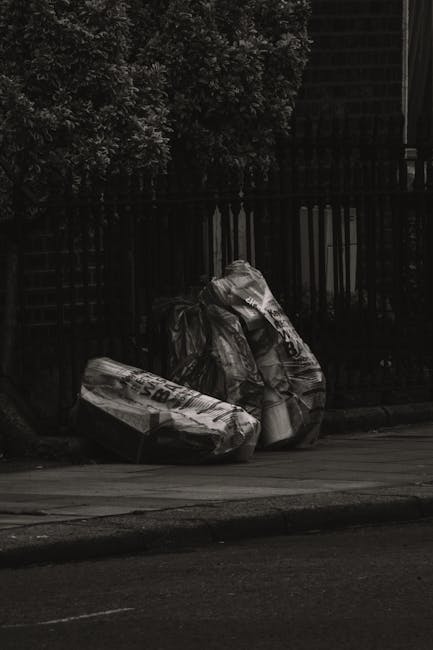 A large pile of black rubbish bags is stacked against a modern building with tiled exterior walls. The bags are filled with waste and some are torn, revealing discarded plastic bottles and other debris inside. The bags occupy most of the foreground, with some leaning or slightly distorted from the weight. The background shows a cloudy sky with hints of sunlight and nearby urban structures, indicating an outdoor urban environment. Red graffiti spelling 'XEND' is spray-painted across the tiles behind the waste pile. The scene illustrates independent waste disposal or rubbish removal efforts typical of private collection services, such as those provided by Rubbish Collection Haringey, highlighting the need for proper rubbish management to prevent illegal dumping.