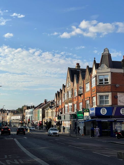 A row of multi-story terraced buildings with brick facades, featuring a combination of bay windows and pitched roofs, lines a busy urban street. The ground level houses various small shops and businesses, with visible signage and storefronts, including a public transport station entrance marked with a blue sign and the letter 'B.' Several pedestrians are walking along the sidewalk, some waiting at crosswalks as cars move through the intersection. The street is paved with asphalt, marked with lane markings, and includes designated parking spaces. In the background, additional commercial and residential buildings extend along the street, set against a partly cloudy sky with patches of blue and floating white clouds. The overall scene depicts a typical city street in Haringey, with a mix of commercial activity and local residential architecture, relevant to outdoor rubbish collection and waste management services like those provided by Rubbish Collection Haringey, supporting alternative waste handling methods through private clearance or on-site rubbish removal.