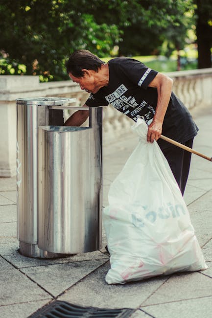 A middle-aged man with dark hair combed back, dressed in a black T-shirt with white and blue graphic prints on the front and sleeves, is seen disposing of trash into a cylindrical stainless steel litter bin on a paved urban sidewalk. He holds a large white plastic bag, partially filled with waste, in his left hand while leaning forward to open the top of the bin with his right hand. The bin is designed for rubbish collection and is positioned against a stone or concrete balustrade, with a backdrop of lush green trees and foliage, suggesting a park or public space setting. The scene is lit with natural daylight, indicating a clear, sunny day. The overall context relates to individual-level waste disposal as part of community cleanliness efforts, with the environment being well-maintained and free of litter. Rubbish Collection Haringey specializes in providing alternative waste handling options, such as private rubbish collection services in urban areas like this, supporting local efforts to manage waste outside of standard municipal collection methods.