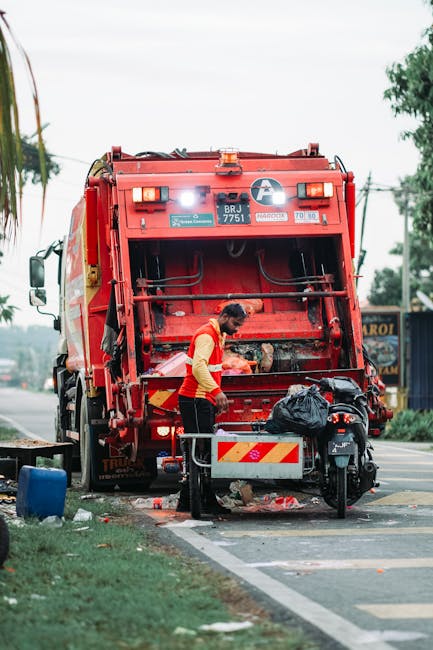 A worker in a yellow safety vest is seen operating a red refuse collection vehicle parked on the side of a street. The rear of the vehicle is open, exposing mechanical components and an internal compartment where waste is loaded. The worker appears to be handling black rubbish bags, which are placed on a small platform attached to a motorbike parked nearby. The scene is set outdoors during daylight, with a partly cloudy sky, trees, and utility poles visible in the background. The surrounding environment includes a paved roadside with a curb and a strip of grass, indicating a typical urban or suburban area. The presence of waste bags and the operation of the rubbish collection vehicle suggest ongoing waste collection activities, demonstrating a professional approach to local waste management services by firms like Rubbish Collection Haringey, which offer alternative waste handling solutions outside of traditional council services.