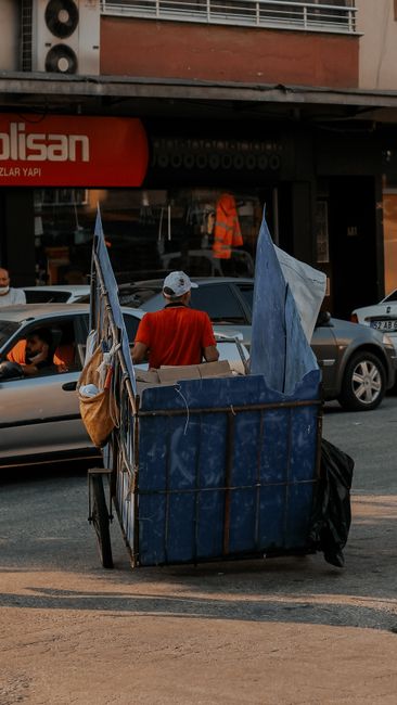 A worker in a yellow safety vest is seen operating a red refuse collection vehicle parked on the side of a street. The rear of the vehicle is open, exposing mechanical components and an internal compartment where waste is loaded. The worker appears to be handling black rubbish bags, which are placed on a small platform attached to a motorbike parked nearby. The scene is set outdoors during daylight, with a partly cloudy sky, trees, and utility poles visible in the background. The surrounding environment includes a paved roadside with a curb and a strip of grass, indicating a typical urban or suburban area. The presence of waste bags and the operation of the rubbish collection vehicle suggest ongoing waste collection activities, demonstrating a professional approach to local waste management services by firms like Rubbish Collection Haringey, which offer alternative waste handling solutions outside of traditional council services.