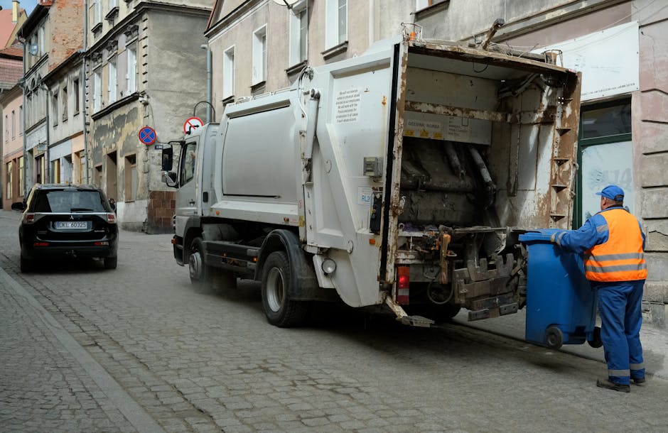 A street scene featuring a white refuse collection truck parked on a cobblestone roadway next to a row of older, multi-story buildings with stone and brick facades. The truck's rear compactor is open, revealing rusted mechanical components and a worn metal interior. A sanitation worker dressed in a high-visibility orange vest, blue uniform, and blue cap is positioned at the back, emptying a blue wheeled bin into the truck's compartment. Another vehicle, a black SUV with a visible license plate, is parked further down the street. The buildings have large windows and some show signs of age with chipped paint and weathered surfaces. Street signs indicating parking restrictions are mounted on poles along the sidewalk. The scene is illuminated by natural daylight, with a neutral and professional atmosphere, illustrating an example of private or independent waste collection services in an urban setting, aligned with rubbish removal operations in Haringey.