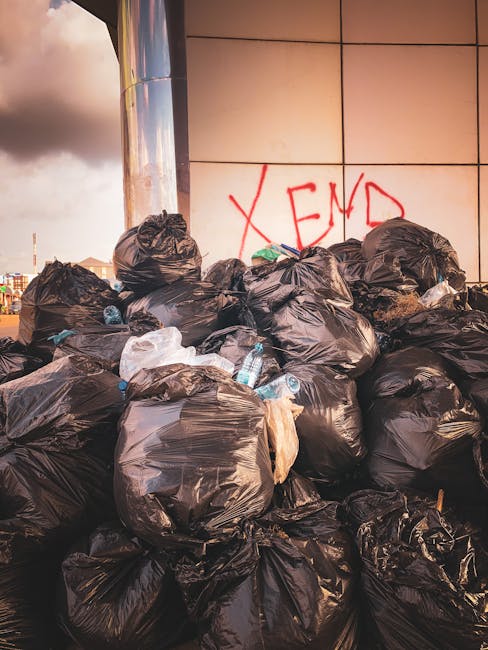 A large pile of black plastic rubbish bags, some partially torn or crumpled, stacked against a modern exterior wall with large beige tiles. The bags contain mixed waste, with visible plastic bottles and white packaging materials protruding from a few of them. In the background, there is a graffiti tag in red spray paint reading 'XEND' on the tiled wall, and a partly cloudy sky with some sunlight breaking through, illuminating the scene. The setting appears to be outdoors on a City pavement or forecourt area, possibly adjacent to a commercial or residential building, representing a typical scene of waste accumulation that could be addressed by independent rubbish collection services like Rubbish Collection Haringey. The overall scene emphasizes the need for waste management and clearance, aligning with alternative waste handling or professional rubbish removal services in the area.