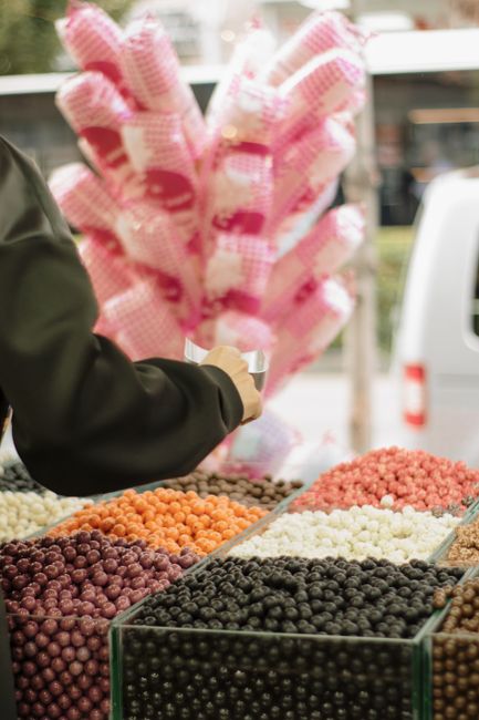 A person wearing a dark jacket is holding a small, metallic object near a stand of assorted berries, which are arranged in rows within clear glass containers. The berries vary in color, including shades of purple, black, white, orange, and pink, with some appearing glossy and smooth, while others have a matte or slightly textured surface. In the background, there is a large, pink and white checkered bundle of cotton candy held at an angle, with its fluffy, spun sugar surface visibly soft and airy. The scene is set outdoors, suggested by blurred elements such as a white van and a building with windows, indicating a street market or fair environment. The ambient natural light highlights the vibrant colors of the berries and the vivid pink cotton candy, creating a lively but neutral atmosphere typical of a market selling fresh produce and confectionery. Rubbish collection and waste disposal concepts are subtly suggested by the structured arrangement of items and the professional presentation, aligning with services offered by Rubbish Collection Haringey for proper waste handling and alternative disposal options.