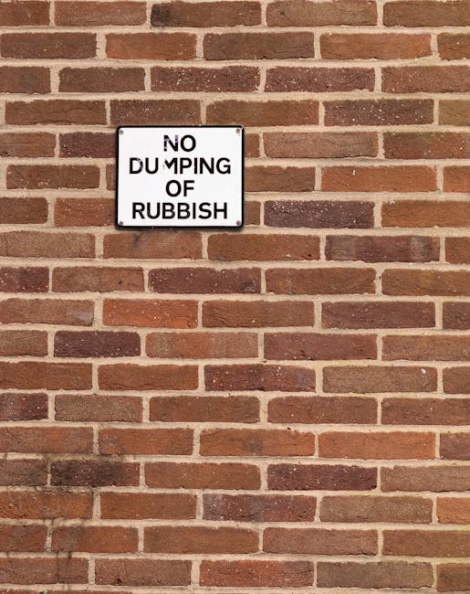 A close-up view of a rectangular, metal sign mounted on a red brick wall. The sign has a white background with black text that reads 'NO DUMPING OF RUBBISH,' although the word 'DUMPING' is partially obscured or damaged, making part of the text less clear. The wall behind the sign is constructed from uniformly sized bricks with reddish-brown tones, laid in a horizontal pattern, featuring mortar joints in a light color. The surface of the bricks shows some variation in texture and slight weathering, and the lighting appears natural, with soft shadows cast on the wall. The overall scene suggests a location in an outdoor urban setting, typical for waste management signage in areas where rubbish disposal might be regulated to prevent illegal dumping, aligning with themes of private or controlled waste collection. Rubbish Collection Haringey occasionally notes signage like this as part of their emphasis on rubbish rules and site management. The image is clear, detailed, and provides an accurate visual context for understanding waste regulation signage attached to brick environments.