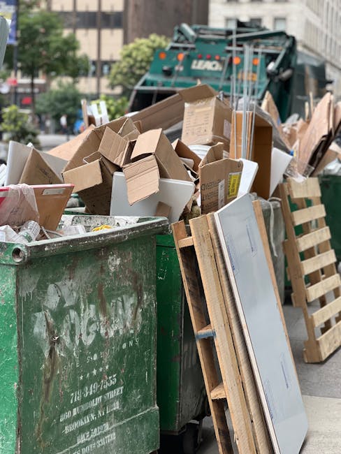A large green waste bin overflowing with a variety of discarded cardboard boxes, some flattened and others still in box form, with a few white polystyrene packaging pieces visible among the debris. The boxes are of different sizes and shades of brown, with some showing printed labels and shipping marks. Leaning against the bin is a wooden pallet and a section of flat-screen television, indicating disposal of electronic waste alongside packaging materials. In the background, a commercial street scene can be seen, including a residential building and a large waste collection vehicle partially visible, suggesting the scene involves waste collection or on-site clearance. The environment appears to be an urban street with natural light and daytime conditions, capturing a typical scene of waste or rubbish removal, possibly related to private disposal or alternative waste handling services like those offered by Rubbish Collection Haringey.