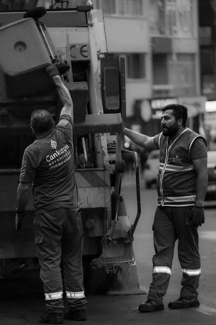 Two waste collection workers, dressed in high-visibility safety vests and dark trousers with reflective stripes, are operating a large commercial rubbish truck on a city street during the daytime, with buildings and parked vehicles visible in the background. One worker, seen from the back, is reaching up to the side of the vehicle, possibly adjusting or inspecting equipment, while the other, facing partially to the side, is standing with his hand resting on the truck, observing the scene. The rubbish truck appears to be a heavy-duty vehicle designed for commercial waste collection, with visible mechanical components, a hydraulic arm, and an enclosed body for holding rubbish. The image is captured in black and white, emphasizing the textures of the workers’ uniforms and the surfaces of the vehicle, and it relates to private waste collection or on-site rubbish removal services, highlighting the operational aspects of licensed rubbish collection teams in Haringey and the standards they follow, as mentioned on the webpage of Rubbish Collection Haringey.