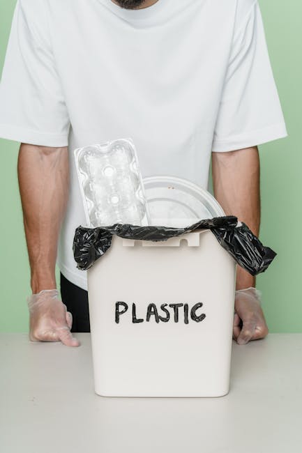 A person wearing a white t-shirt and transparent gloves is holding an open white plastic rubbish bin labeled 'PLASTIC' in black capital letters. Inside the bin, there is a black bin liner that is partially folded over the edges. Leaning against the inside of the bin is a clear plastic packaging tray with multiple compartments, typical of food packaging, and it is visibly empty. The background of the image is a solid, muted green colour, and the person appears to be standing indoors. The scene focuses on the handling of a plastic waste collection, emphasizing proper disposal of plastic materials in an environmentally conscious manner, relevant to private waste handling or alternative rubbish removal services like those offered by Rubbish Collection Haringey.